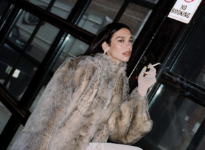 Dua Lipa holding a cigarette outside a dark-fronted building, with a No Smoking sign visible above her.